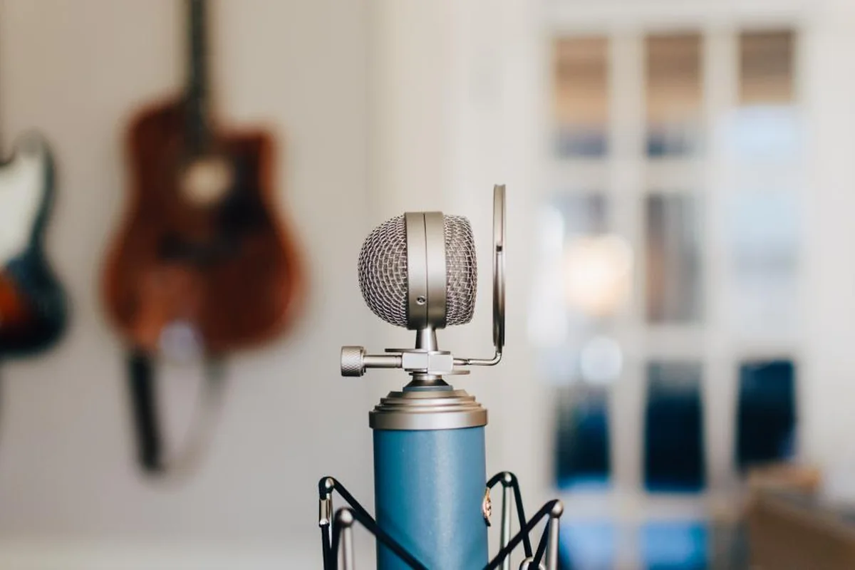 Micrófono de estudio azul y plateado sobre un soporte, con dos guitarras colgadas en la pared desenfocadas al fondo
