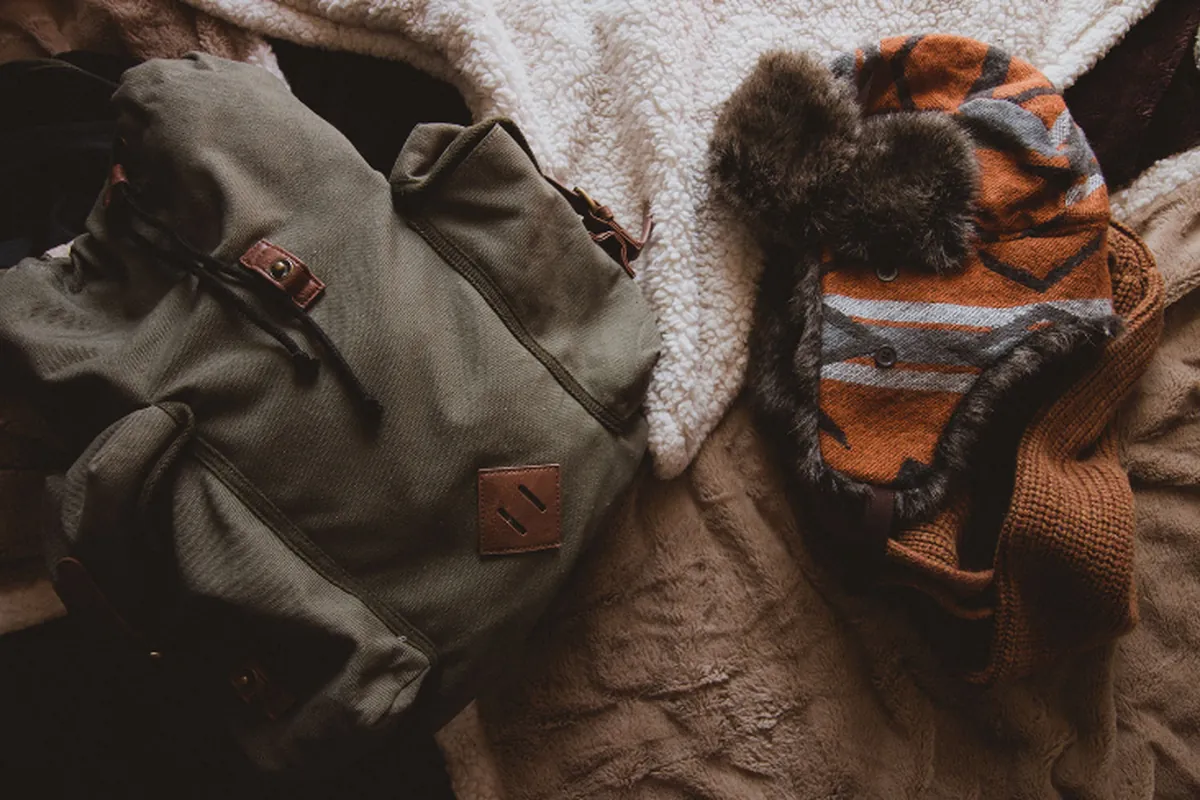 Mochila de lona verde y gorro de lana naranja con forro de pelo sobre una manta clara