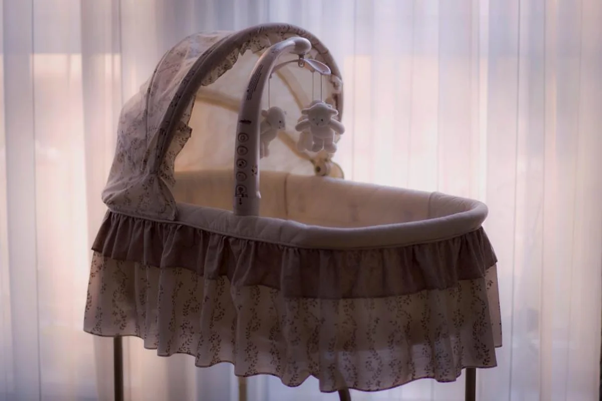 Un moisés con volantes y un móvil de peluches colgando, a contraluz delante de unas cortinas blancas