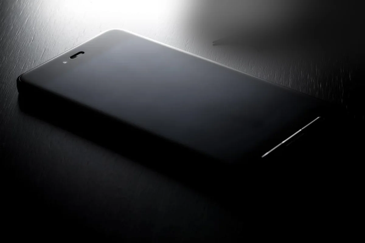 Teléfono móvil con la pantalla apagada sobre una mesa oscura, iluminado por una luz lateral tenue
