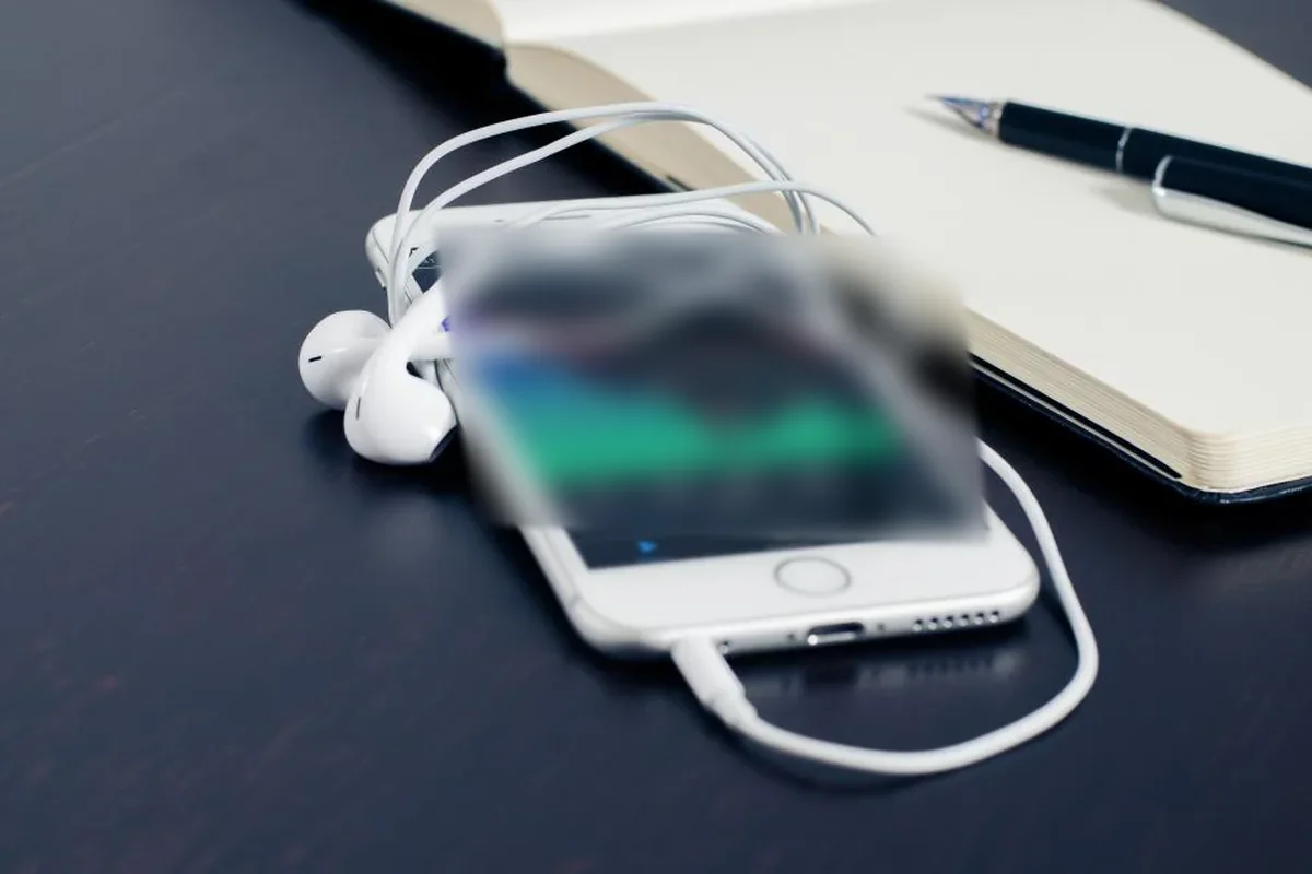 Un móvil con la pantalla encendida y unos auriculares enredados sobre un escritorio oscuro, junto a un cuaderno abierto y un bolígrafo