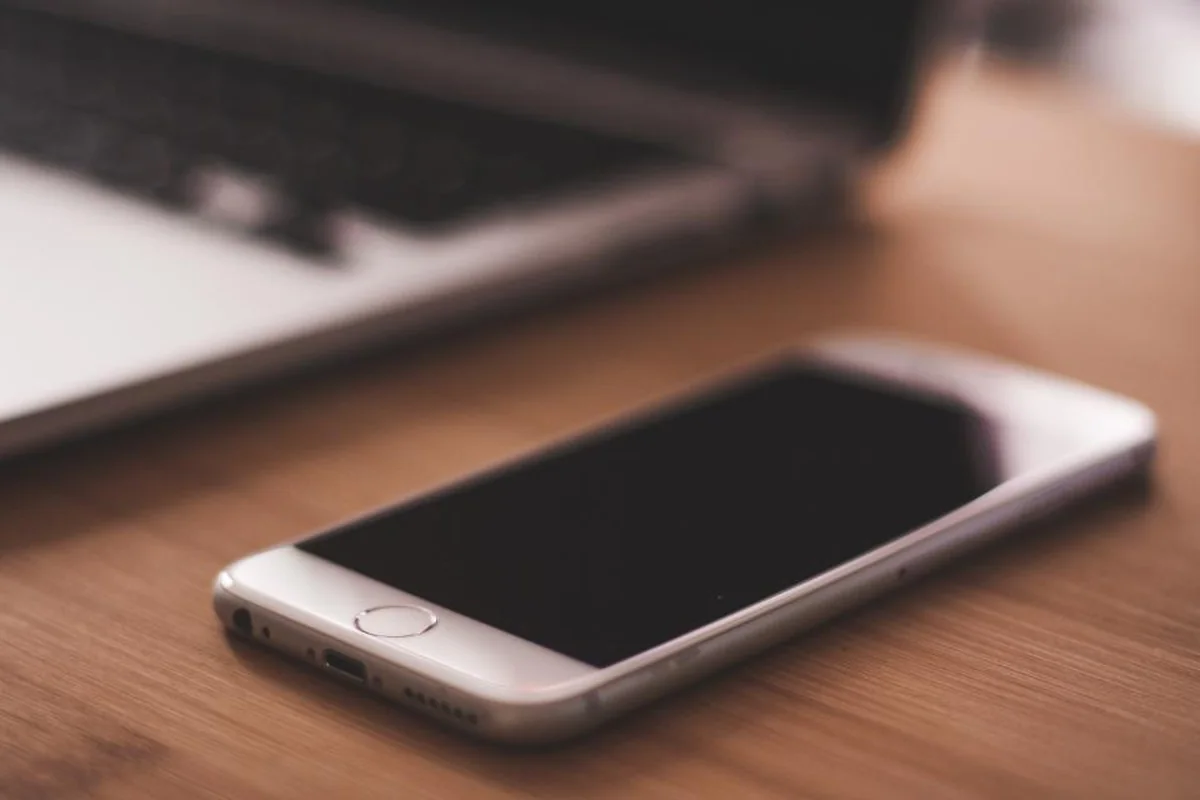 Teléfono móvil con la pantalla apagada sobre una mesa de madera, con un portátil desenfocado al fondo