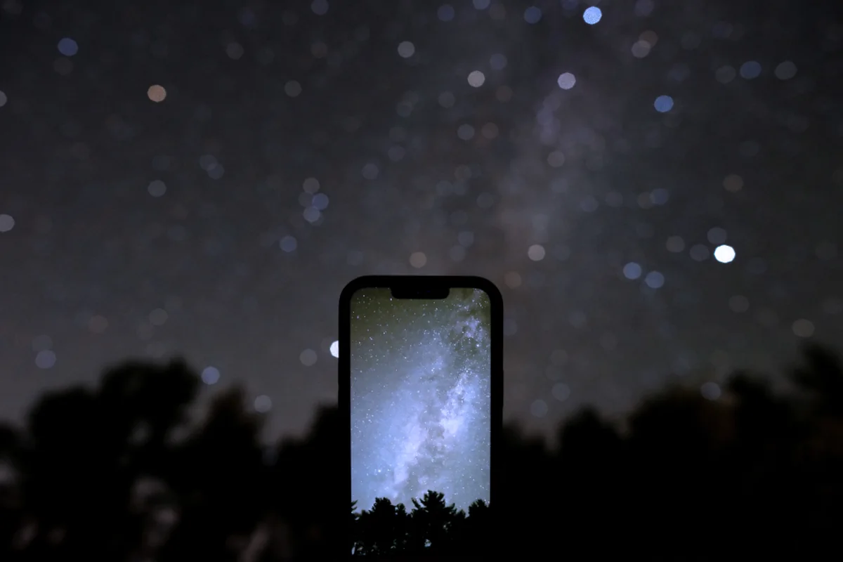Silueta de un móvil sostenido en alto bajo un cielo nocturno lleno de estrellas, con la Vía Láctea reflejada en la pantalla y árboles oscuros al fondo