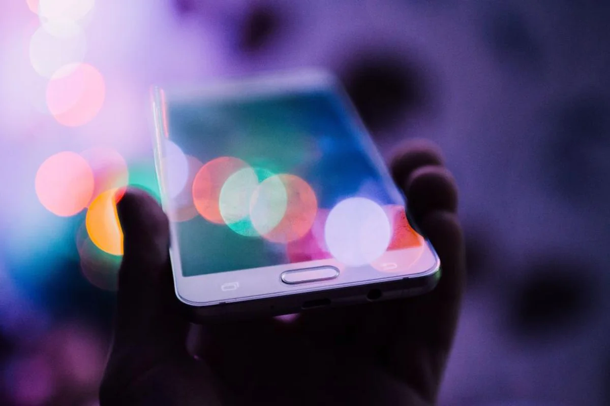 Mano sosteniendo un teléfono móvil en penumbra con luces de colores desenfocadas al fondo