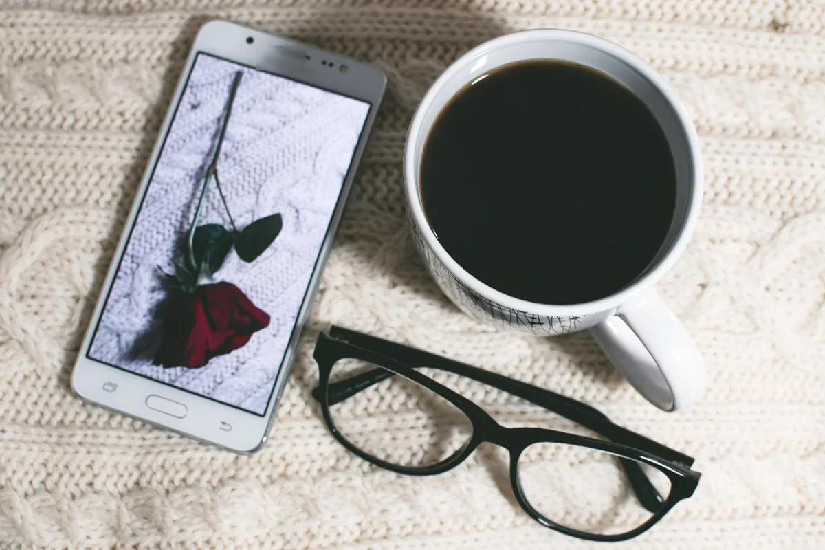 Teléfono móvil con la imagen de una rosa en la pantalla junto a una taza de café y unas gafas sobre una manta de punto