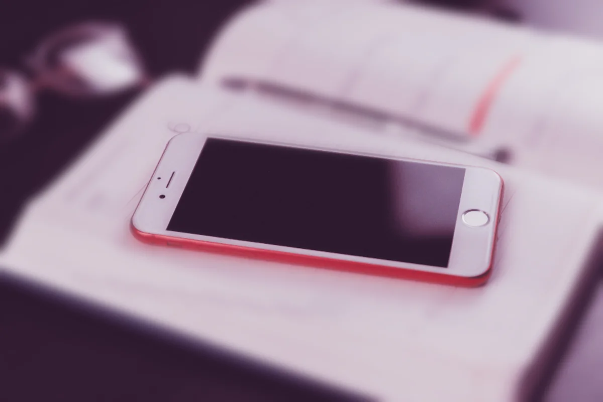 Un teléfono móvil de carcasa roja apoyado sobre una agenda abierta, con un bolígrafo y unas gafas desenfocados al fondo