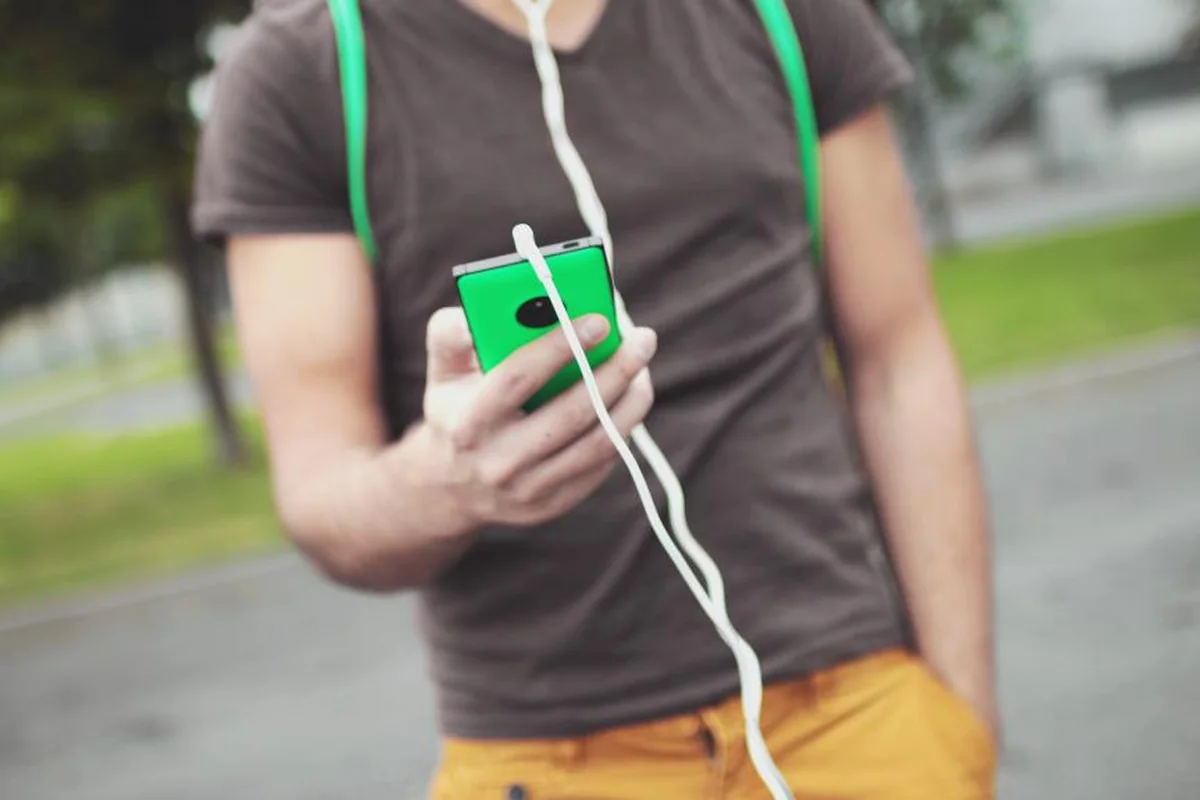 Una persona con camiseta marrón y auriculares con cable sostiene un móvil verde a la altura del pecho, en una calle con árboles desenfocados