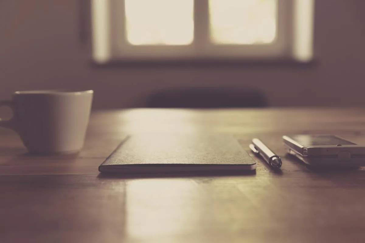 Taza, cuaderno cerrado, bolígrafo y teléfono sobre una mesa de madera iluminada por la luz tenue de una ventana