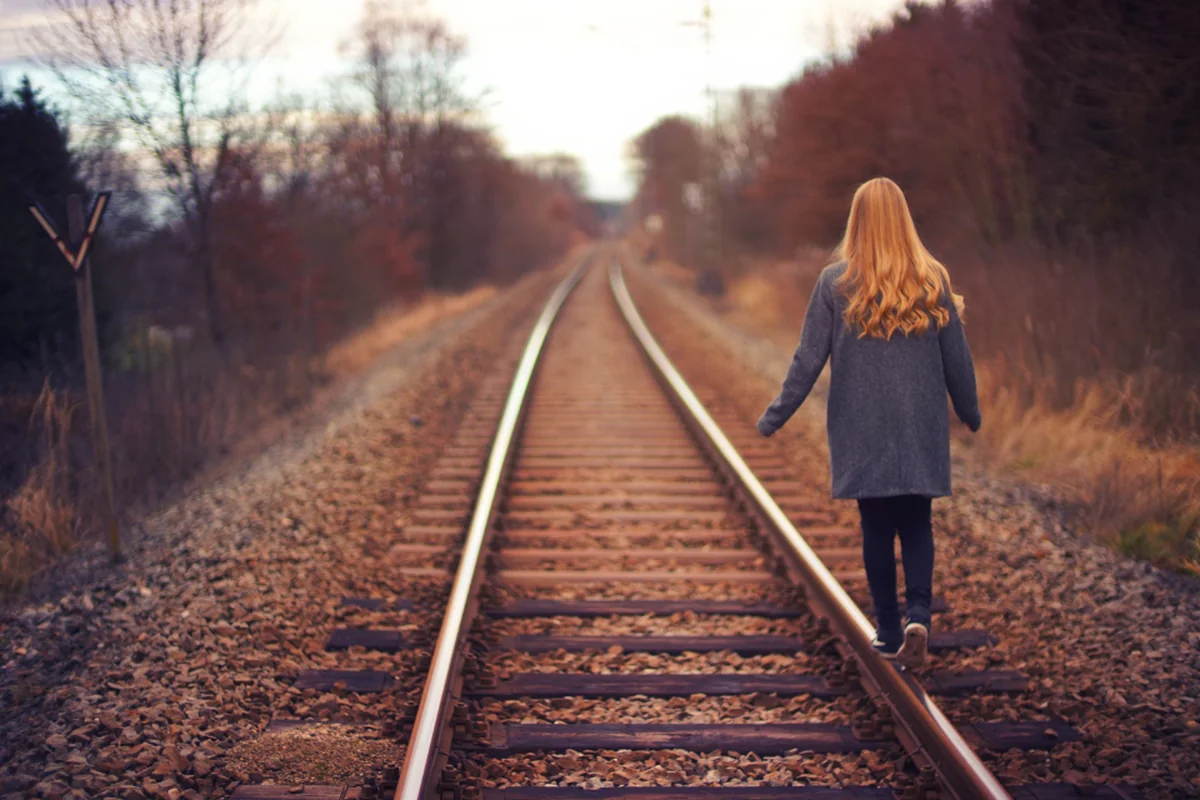 Una mujer de espaldas, con abrigo gris y pelo largo pelirrojo, camina en equilibrio sobre un raíl de una vía de tren rodeada de árboles otoñales