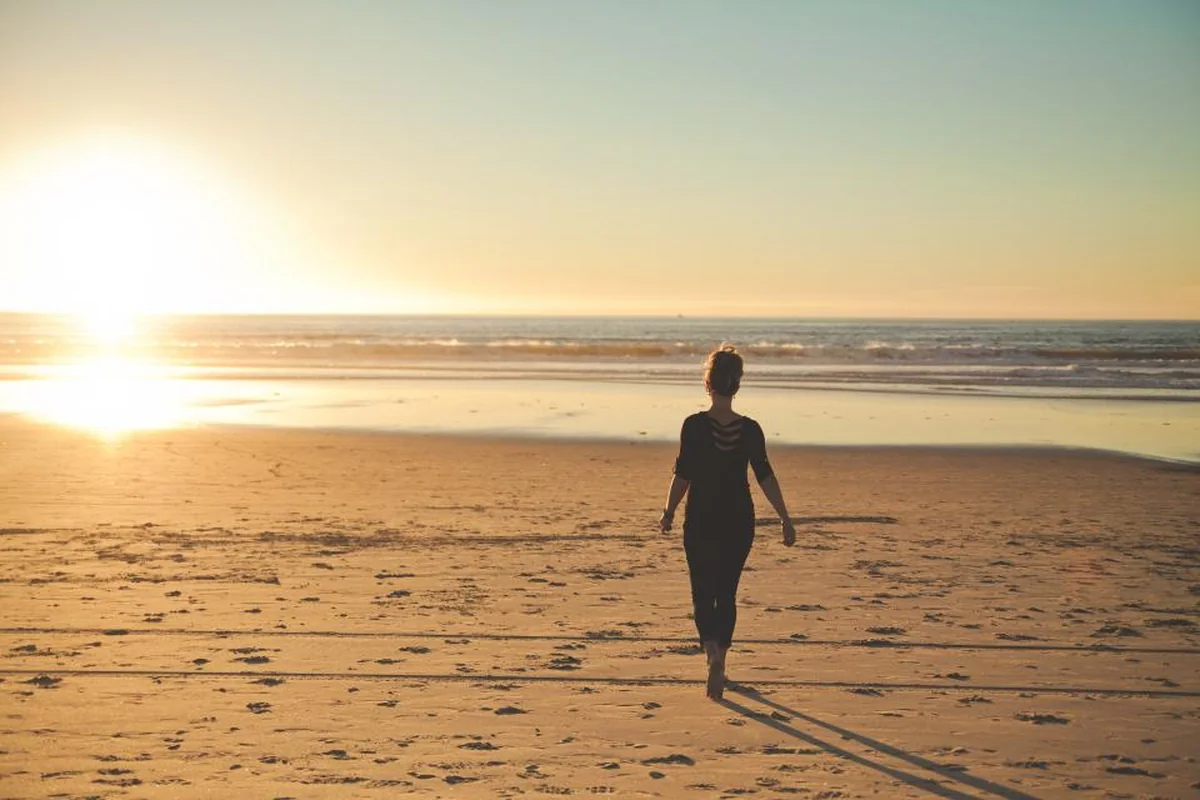 Mujer de espaldas caminando sola por una playa amplia al atardecer, con el sol bajo reflejado en la arena mojada