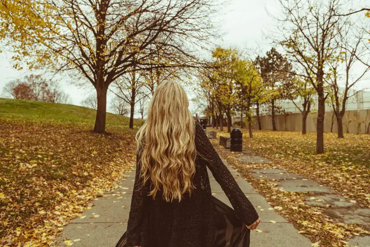 Una mujer de espaldas, con el pelo largo y un abrigo oscuro, camina por un sendero de un parque cubierto de hojas amarillas