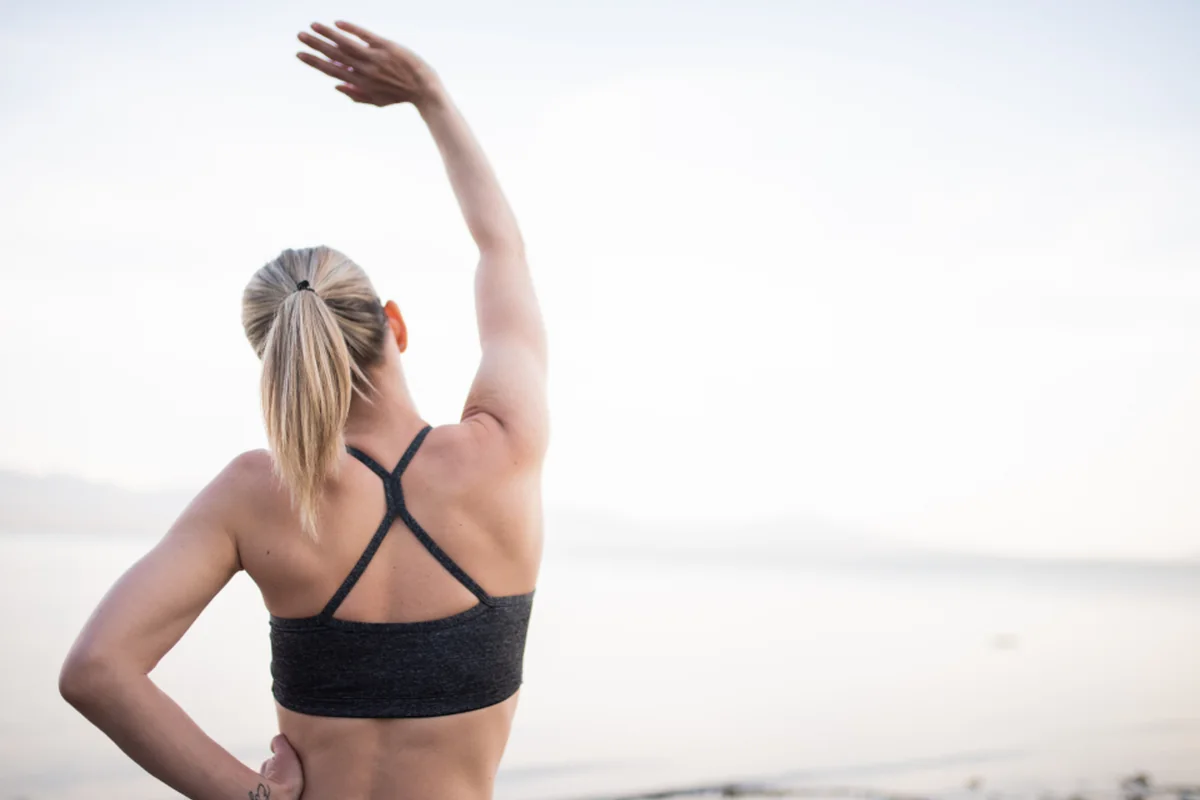 Mujer de espaldas con coleta y ropa deportiva estirando un brazo por encima de la cabeza frente al mar