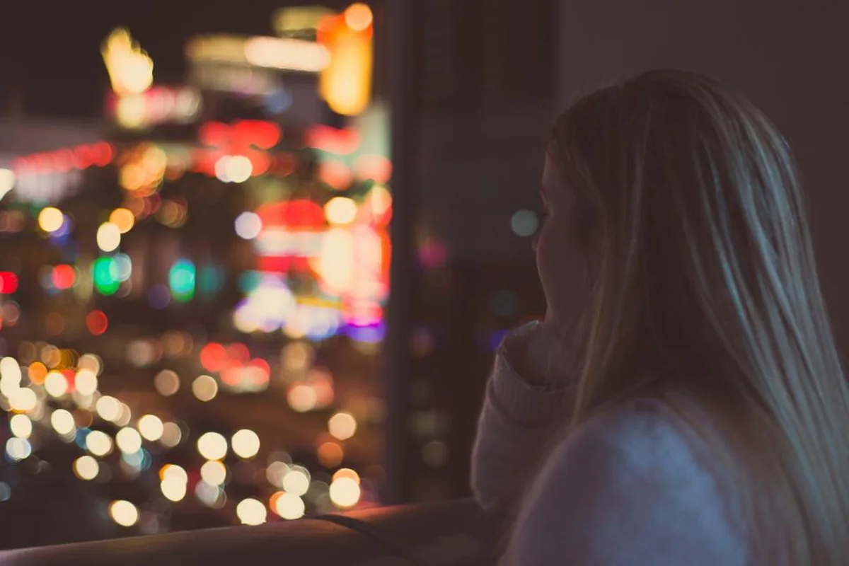 Mujer de espaldas, con el pelo largo, mirando por una ventana las luces desenfocadas de una ciudad de noche