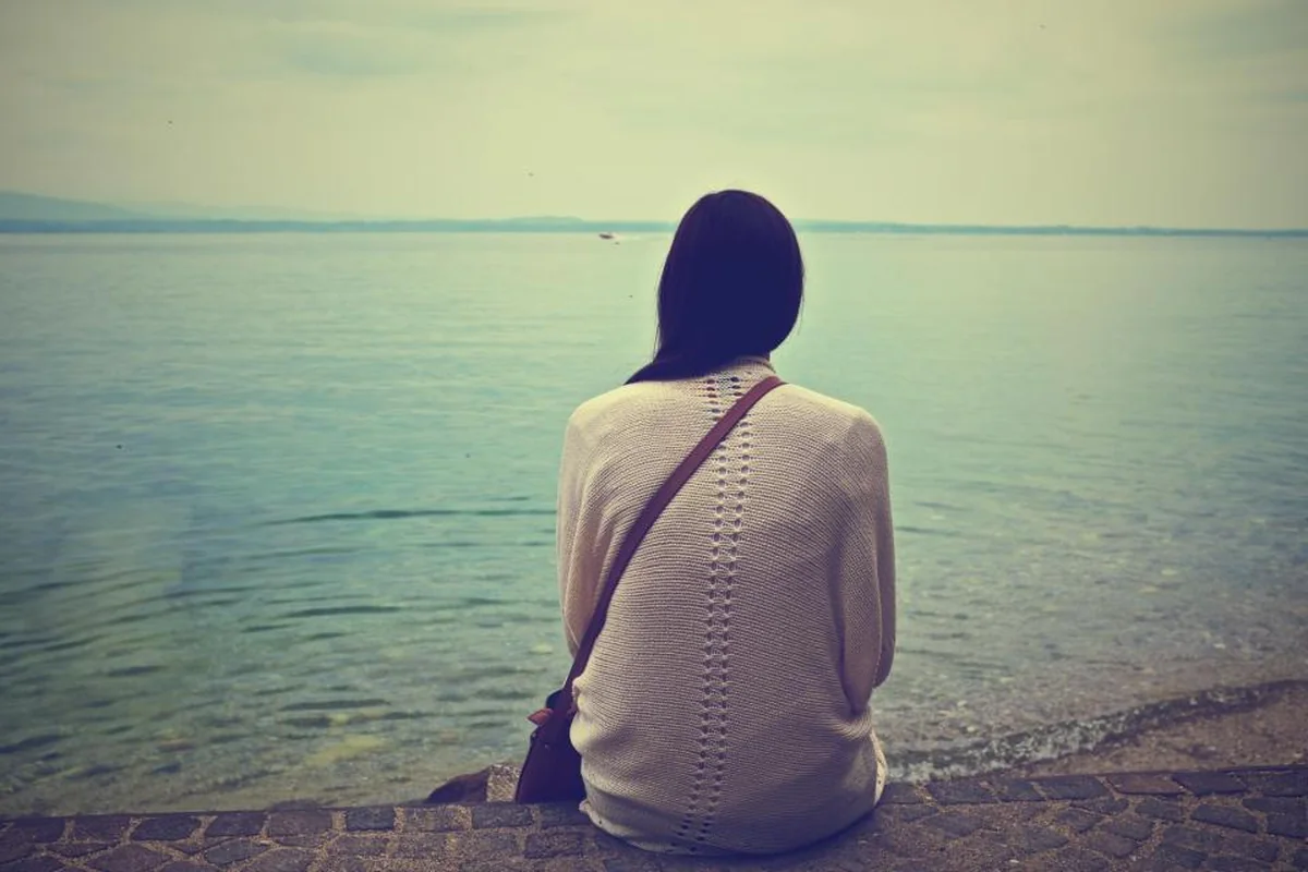 Mujer de espaldas con jersey claro y bolso cruzado, sentada en un muelle de piedra mirando el mar en calma