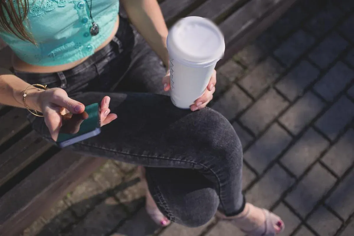 Mujer sentada en un banco de madera, vista desde arriba sin que se le vea la cara, con un vaso de café en una mano y el móvil en la otra