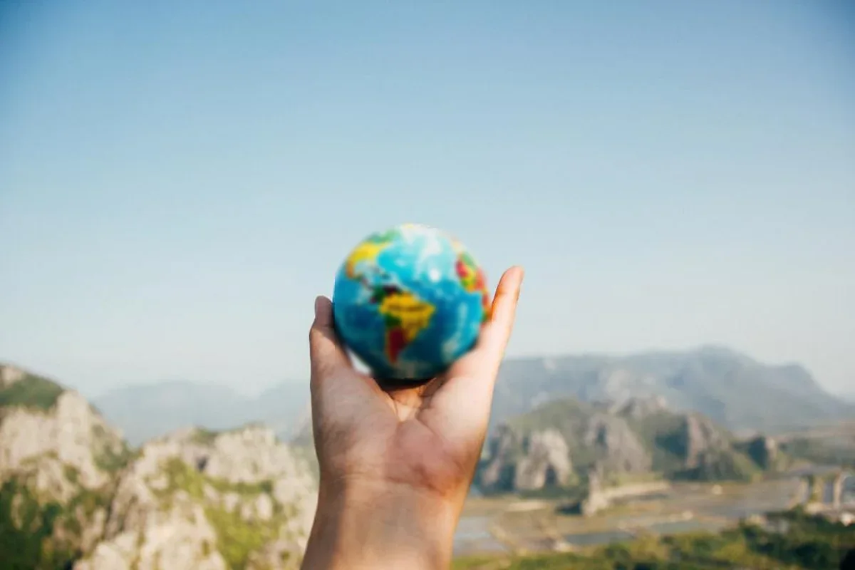 Mano sosteniendo una pequeña bola del mundo frente a un paisaje de montañas desenfocado bajo un cielo despejado