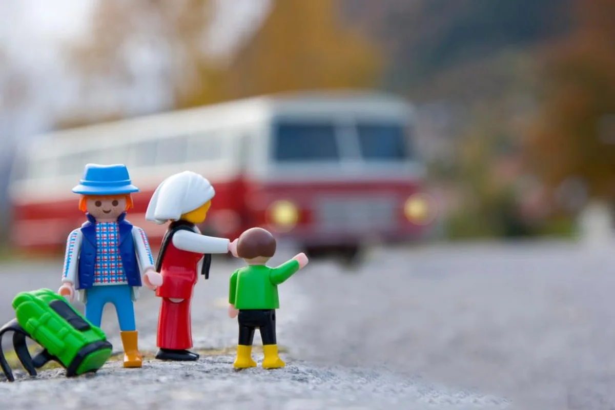 Muñecos de juguete de una familia con mochilas junto a una carretera; el niño señala hacia otro lado mientras pasa un autobús desenfocado