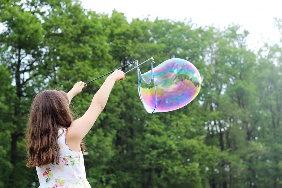 Niña de espaldas con el pelo largo haciendo una gran pompa de jabón con dos varillas en un parque con árboles