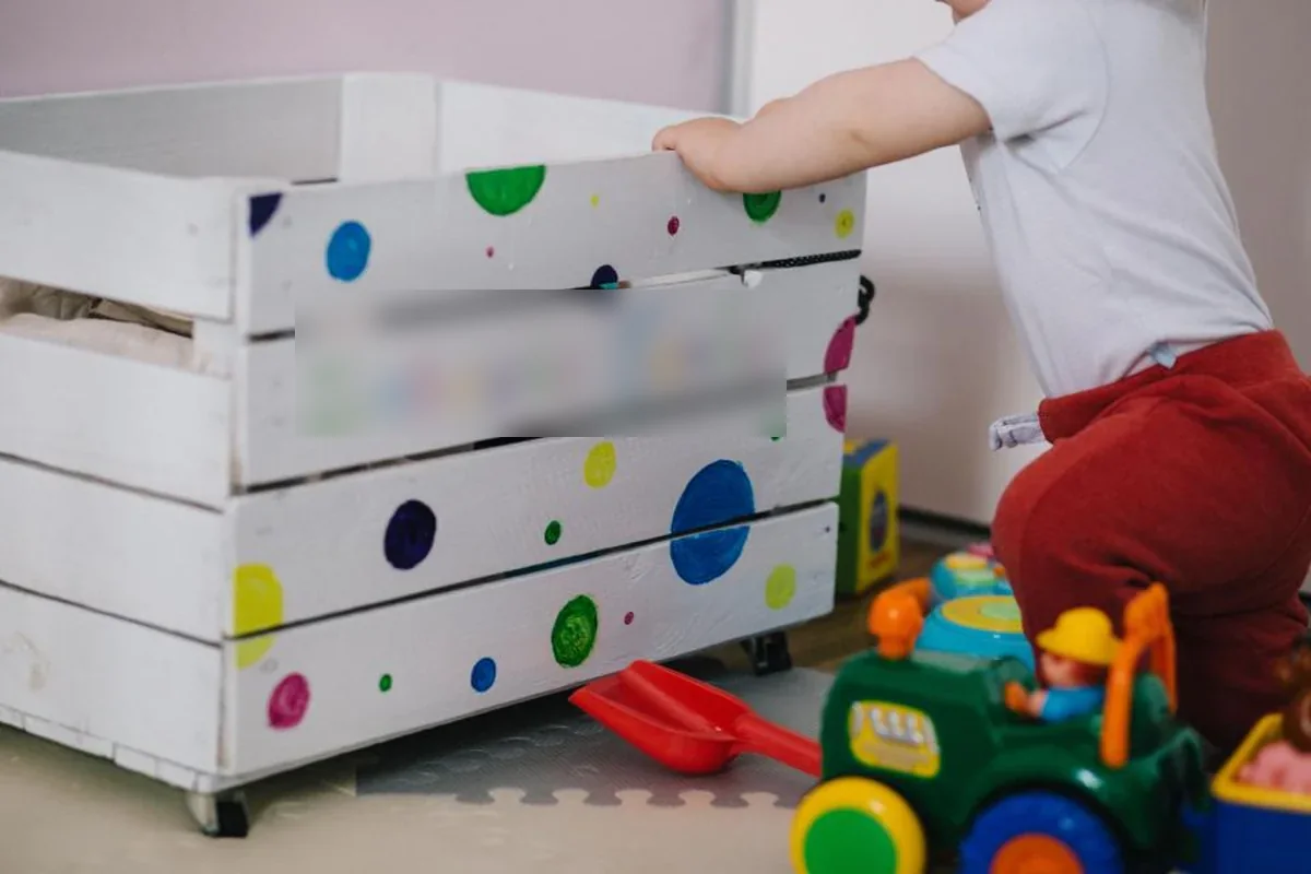 Niño pequeño de espaldas apoyado en una caja de madera blanca con lunares de colores, junto a un tractor de juguete
