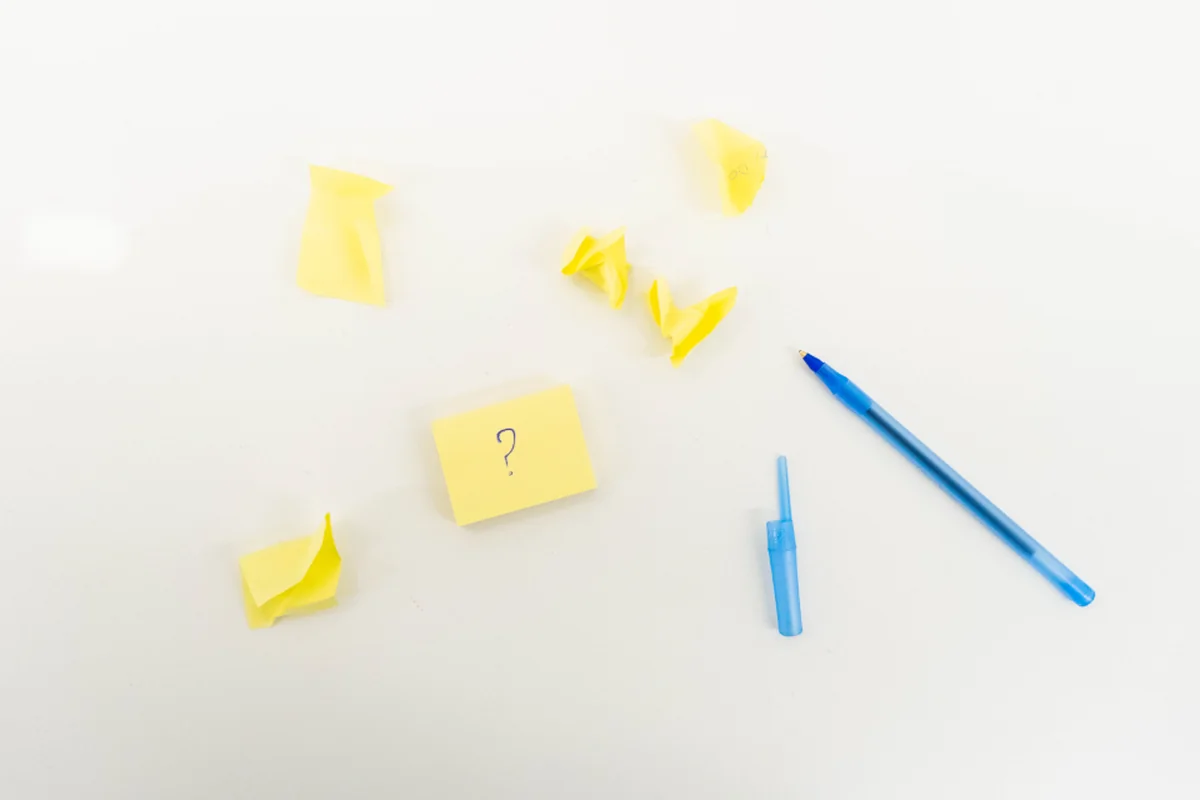Varias notas adhesivas amarillas arrugadas, una con un signo de interrogación dibujado, junto a un bolígrafo azul y su capuchón sobre una mesa blanca