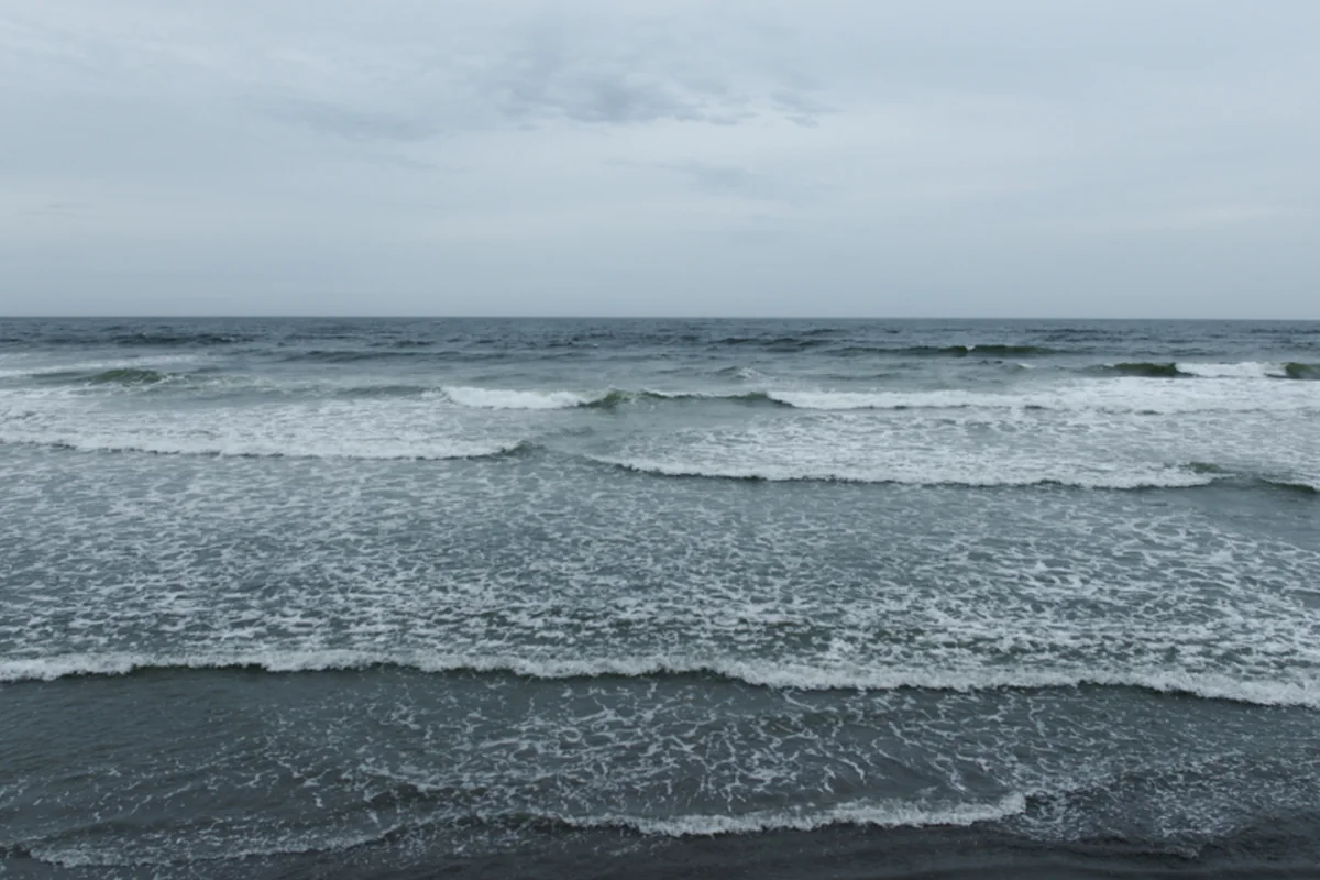 Un mar gris verdoso con varias filas de olas pequeñas rompiendo en la orilla bajo un cielo completamente cubierto