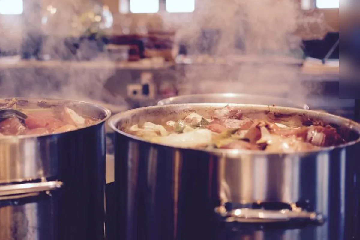 Dos grandes ollas de acero llenas de verduras y carne desprendiendo vapor en una cocina de restaurante con luces cálidas al fondo