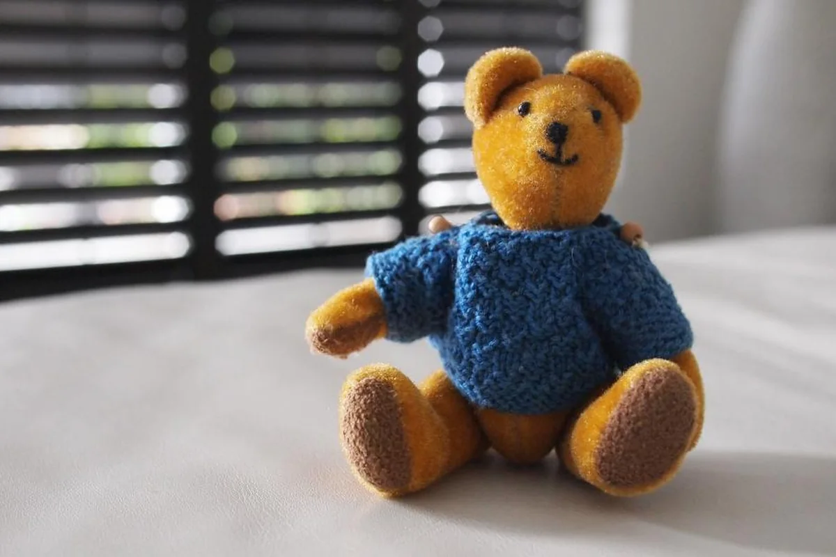 Un osito de peluche con jersey azul de punto sentado sobre una cama junto a una persiana entreabierta