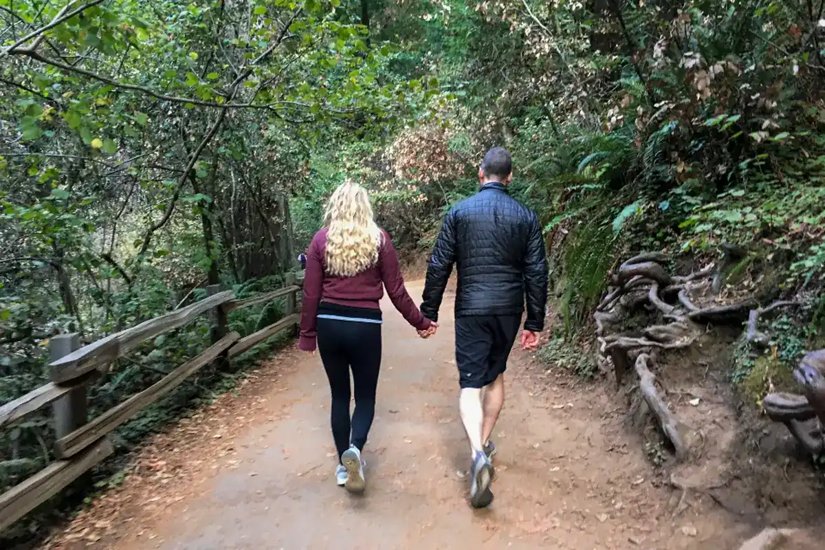 Una pareja de espaldas camina de la mano por un sendero de tierra en un bosque frondoso, junto a una barandilla de madera