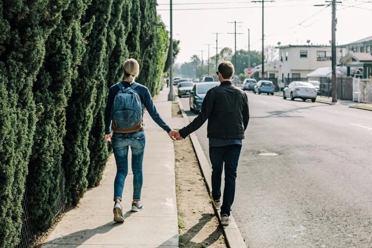 Una pareja joven camina de espaldas por una acera junto a un seto, cogidos de la mano, ella con una mochila vaquera