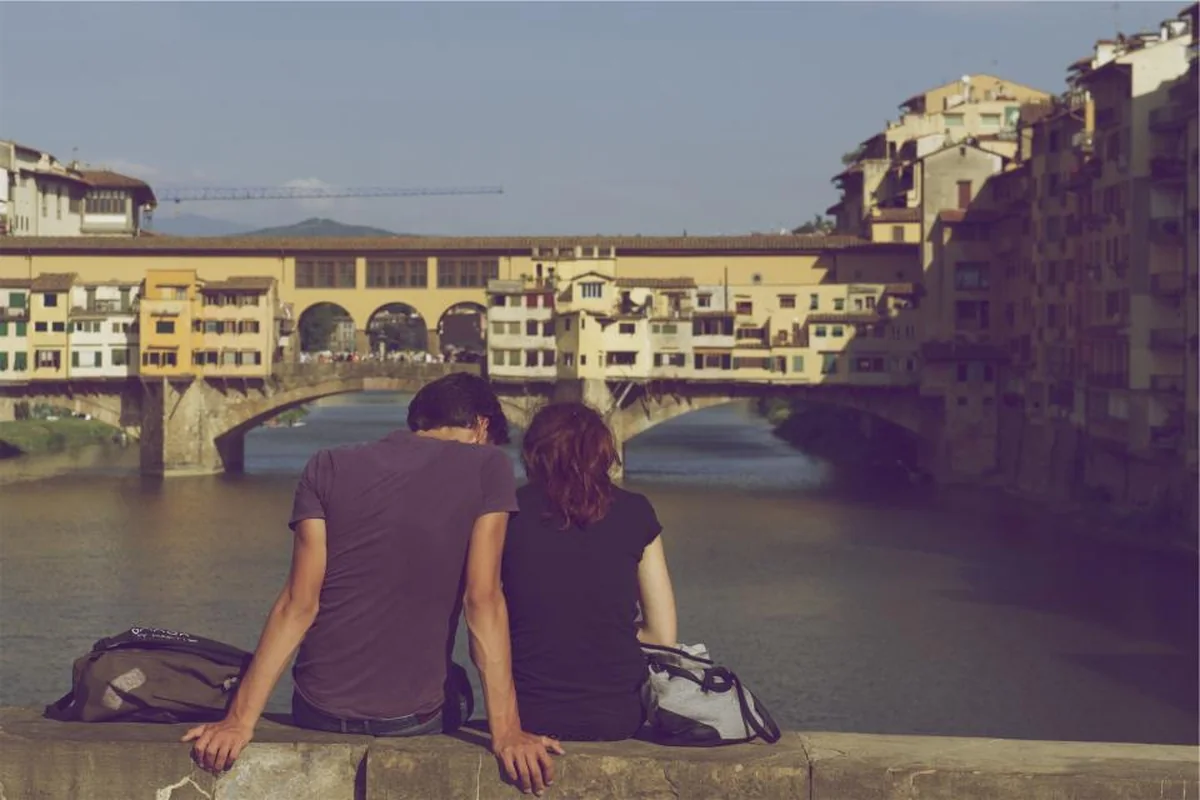 Pareja sentada de espaldas en un muro junto a un río, con un puente y edificios antiguos al fondo