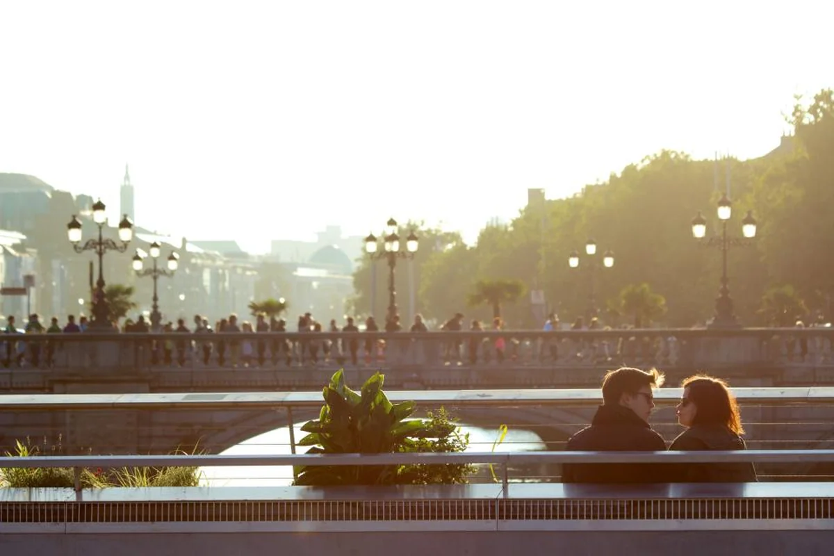 Pareja sentada en un banco junto a un río, mirándose mientras hablan, con un puente y farolas al fondo a contraluz
