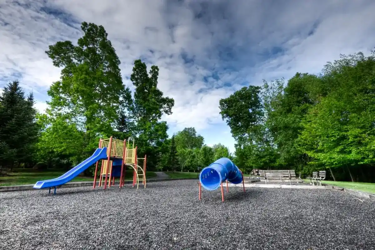 Parque infantil vacío con un tobogán azul y un túnel de plástico sobre suelo de grava, rodeado de árboles
