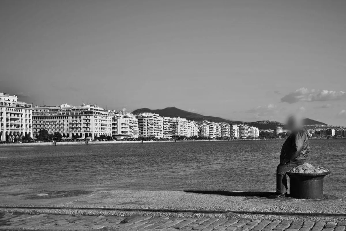 Fotografía en blanco y negro de una persona mayor sentada sobre un noray en un paseo marítimo, mirando al mar, con edificios y montañas al fondo