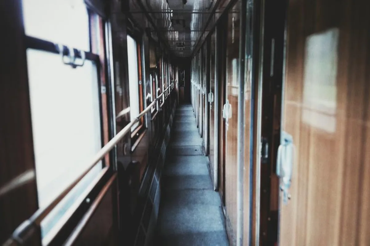 Pasillo estrecho y vacío de un vagón de tren antiguo con paredes de madera, barandilla y ventanas iluminadas a la izquierda
