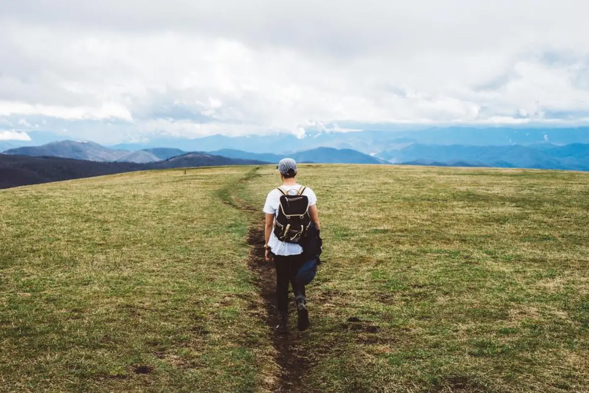 Persona de espaldas con gorra y mochila caminando por un sendero de tierra en un prado verde, con montañas azuladas y cielo nublado al fondo