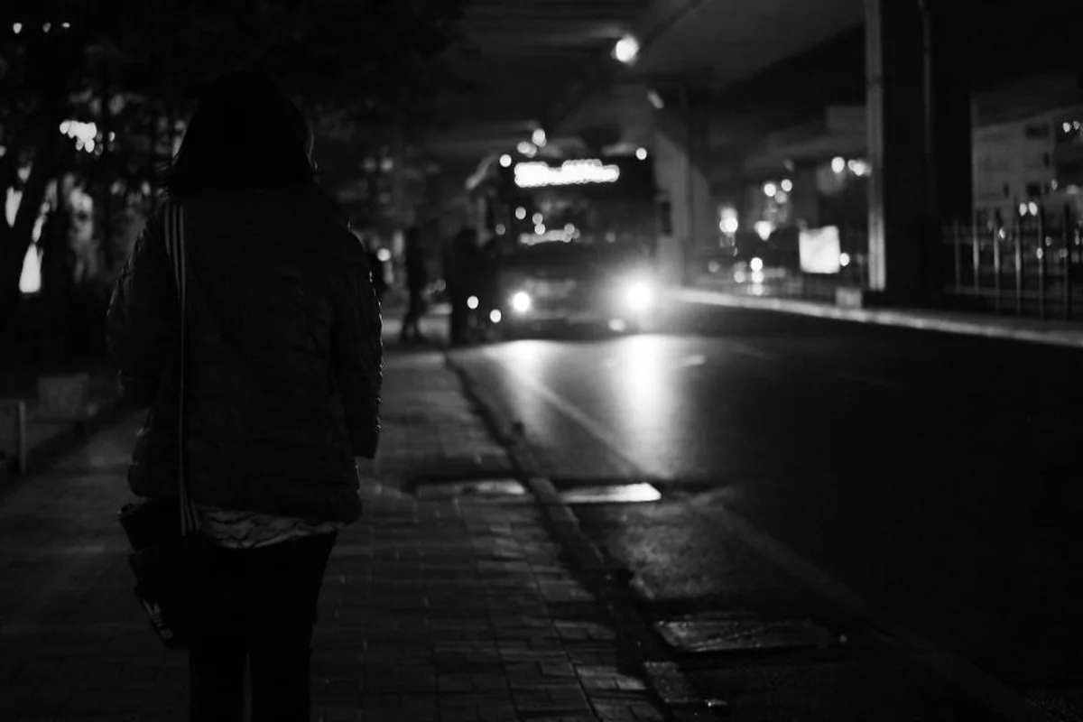 Persona de espaldas con abrigo en una acera de noche mientras se acerca un autobús con los faros encendidos, en blanco y negro