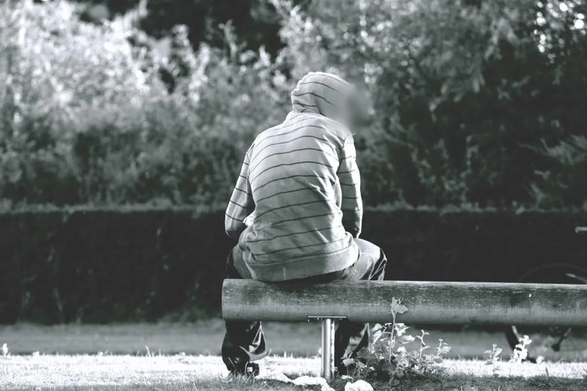 Persona de espaldas con capucha sentada en un banco de madera en un parque, en blanco y negro, frente a un seto y árboles