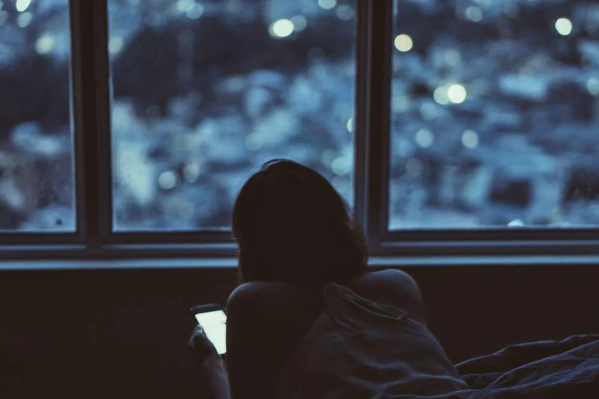 Persona tumbada en la cama, de espaldas, mirando el móvil junto a una ventana de noche