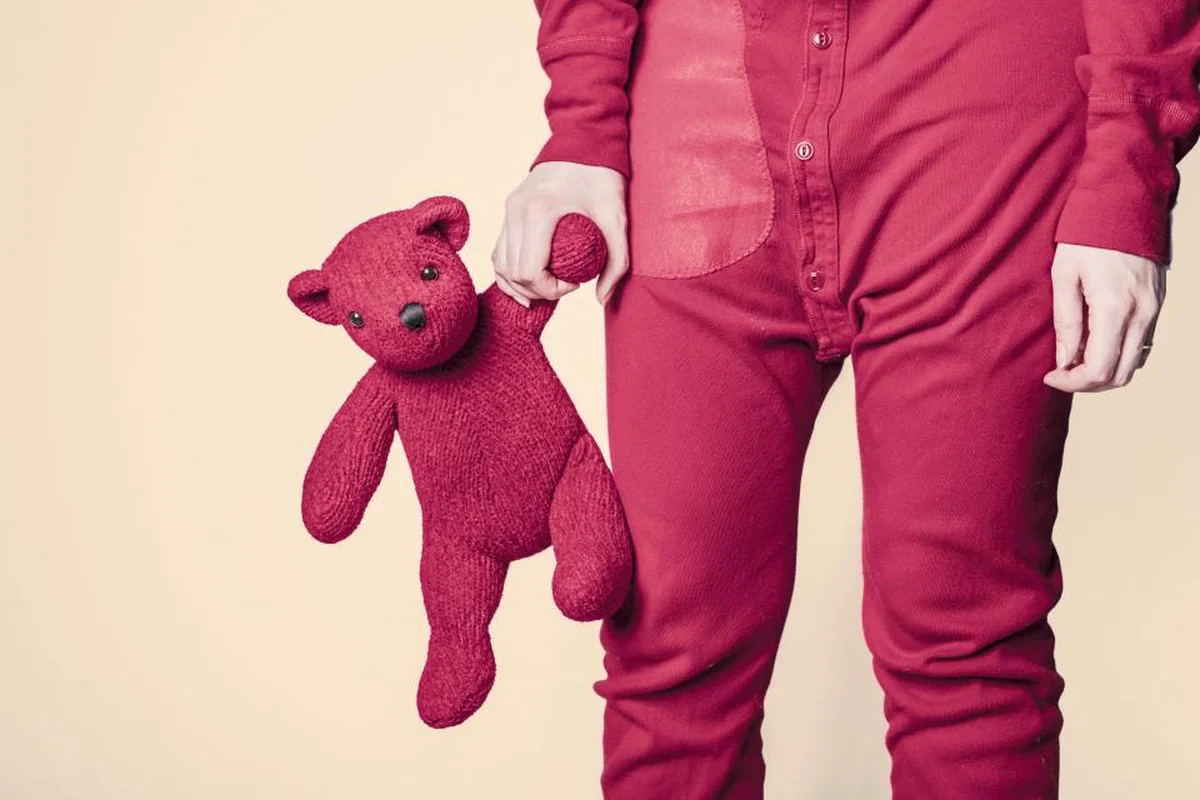 Persona con un pijama rojo de cuerpo entero sujetando de la mano un osito de peluche del mismo color sobre fondo claro