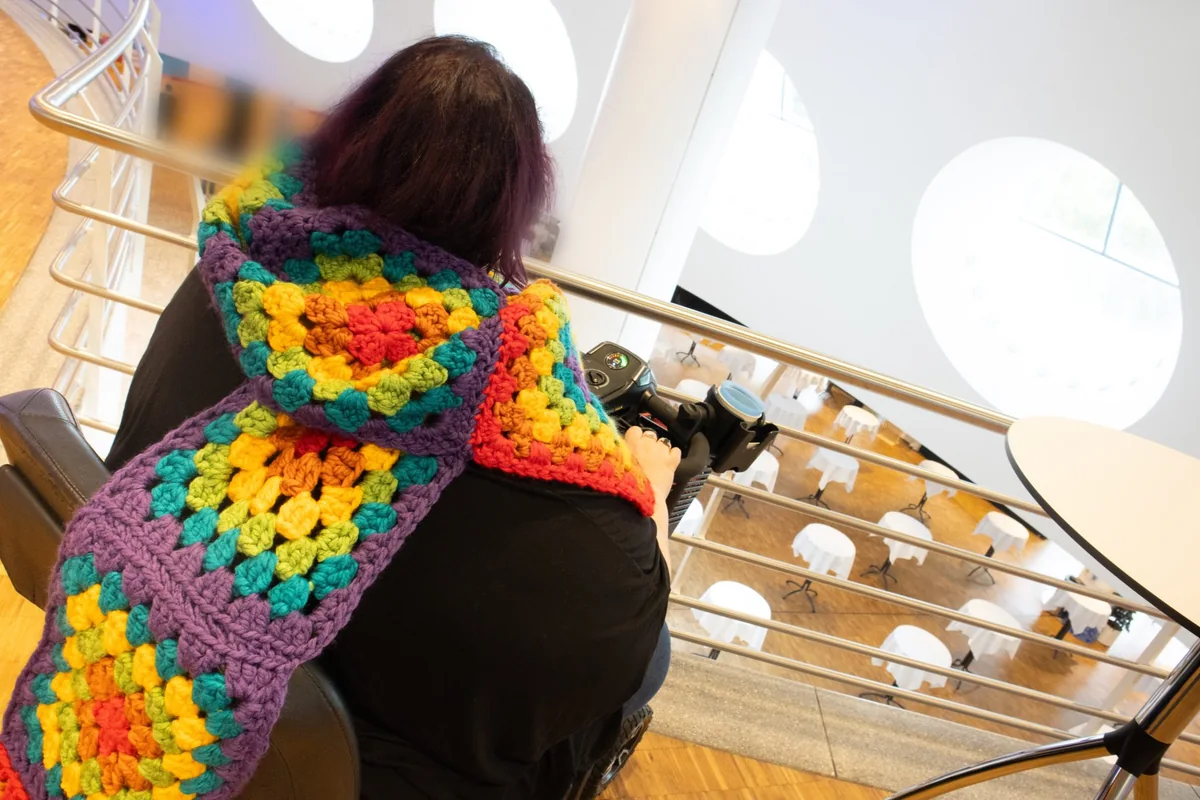 Persona de espaldas en una silla de ruedas eléctrica con una bufanda de ganchillo de colores, junto a la barandilla de un edificio luminoso