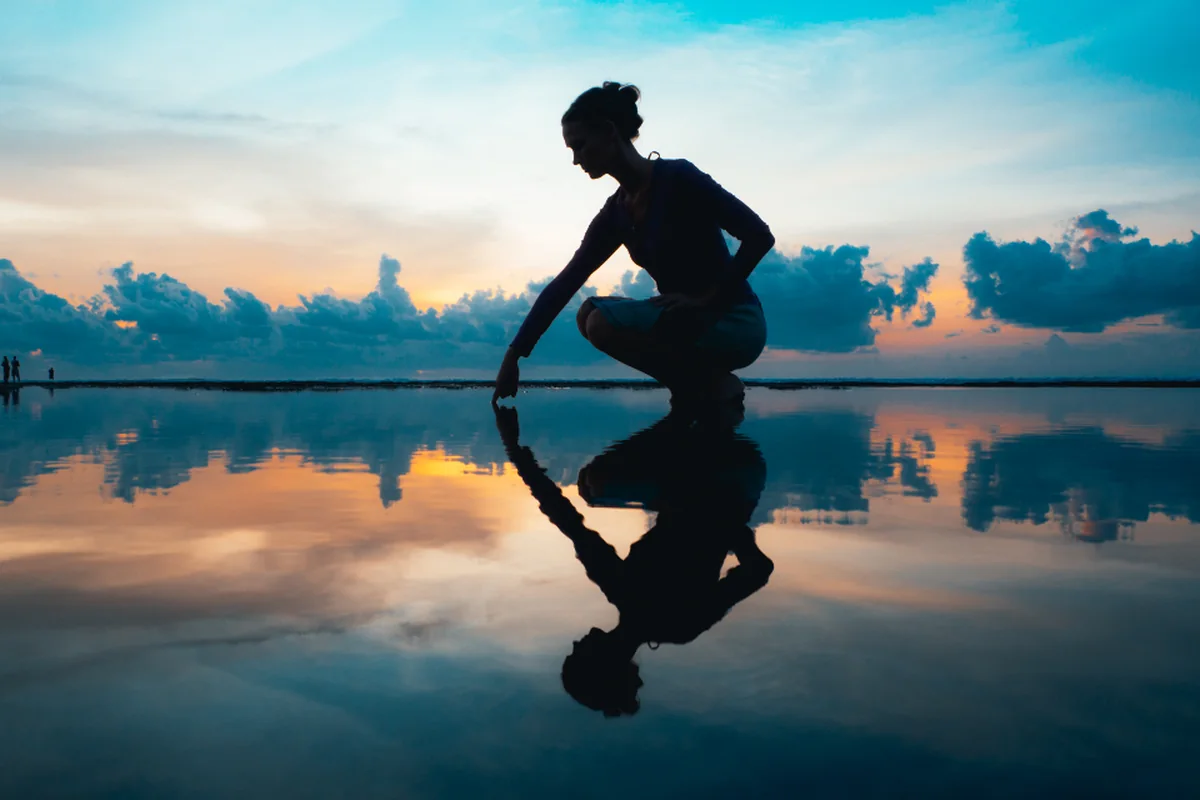 Silueta de una persona agachada tocando el agua en calma, que refleja su figura y las nubes del atardecer