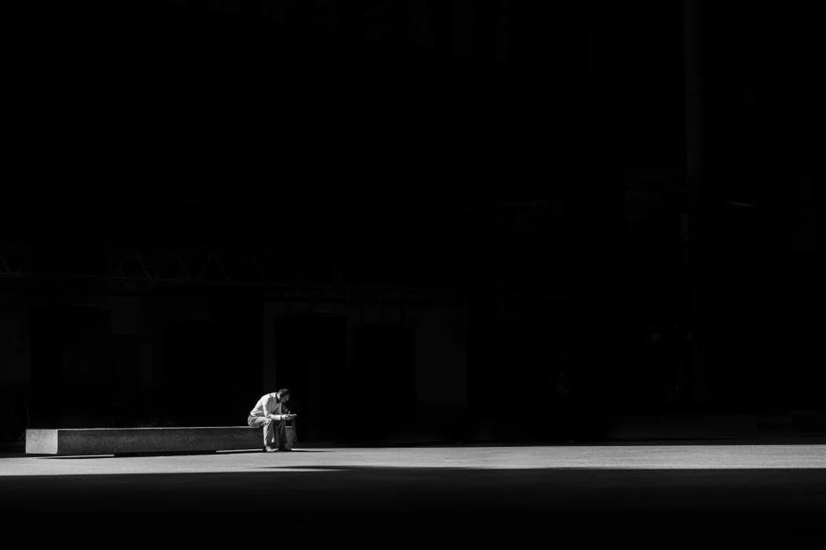 Persona sentada sola en un banco en un espacio amplio y oscuro, en blanco y negro