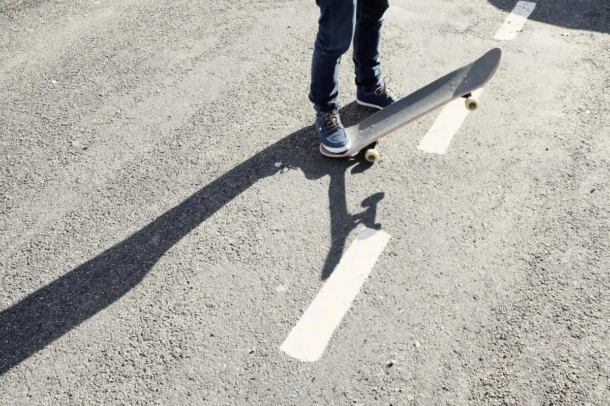 Piernas de una persona con vaqueros y zapatillas pisando un monopatín sobre el asfalto, con su sombra alargada