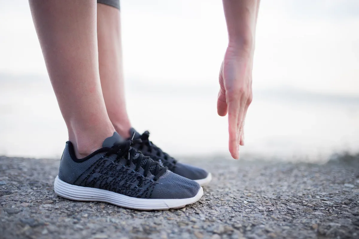 Piernas de una persona con zapatillas deportivas grises estirando sobre el suelo junto al mar, con una mano que baja hacia los pies
