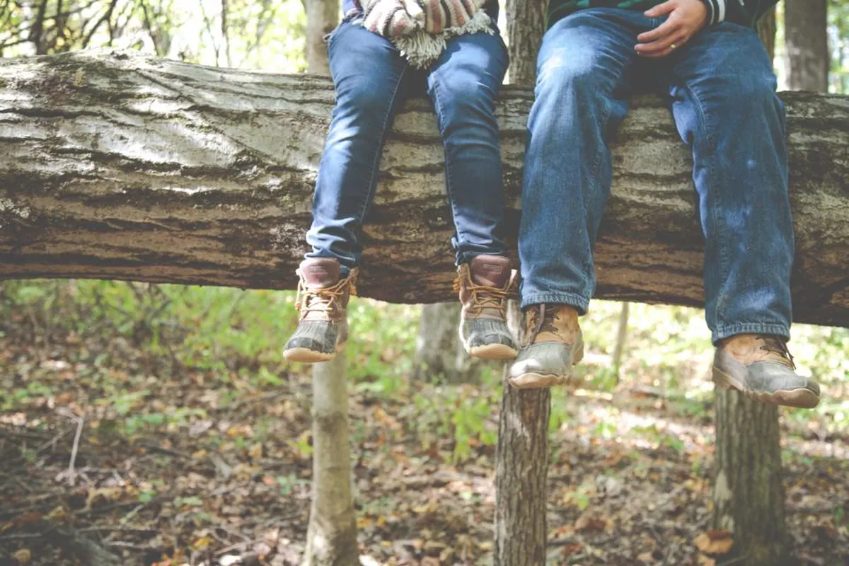 Dos personas sentadas sobre un tronco en un bosque, vistas solo de cintura para abajo con vaqueros y botas
