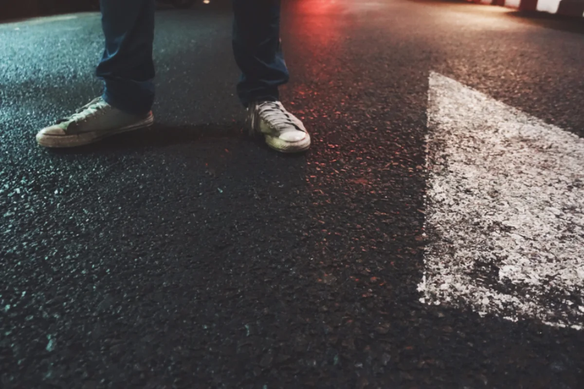 Pies de una persona con vaqueros y zapatillas blancas, parada sobre el asfalto mojado de una calle junto a una flecha pintada en el suelo