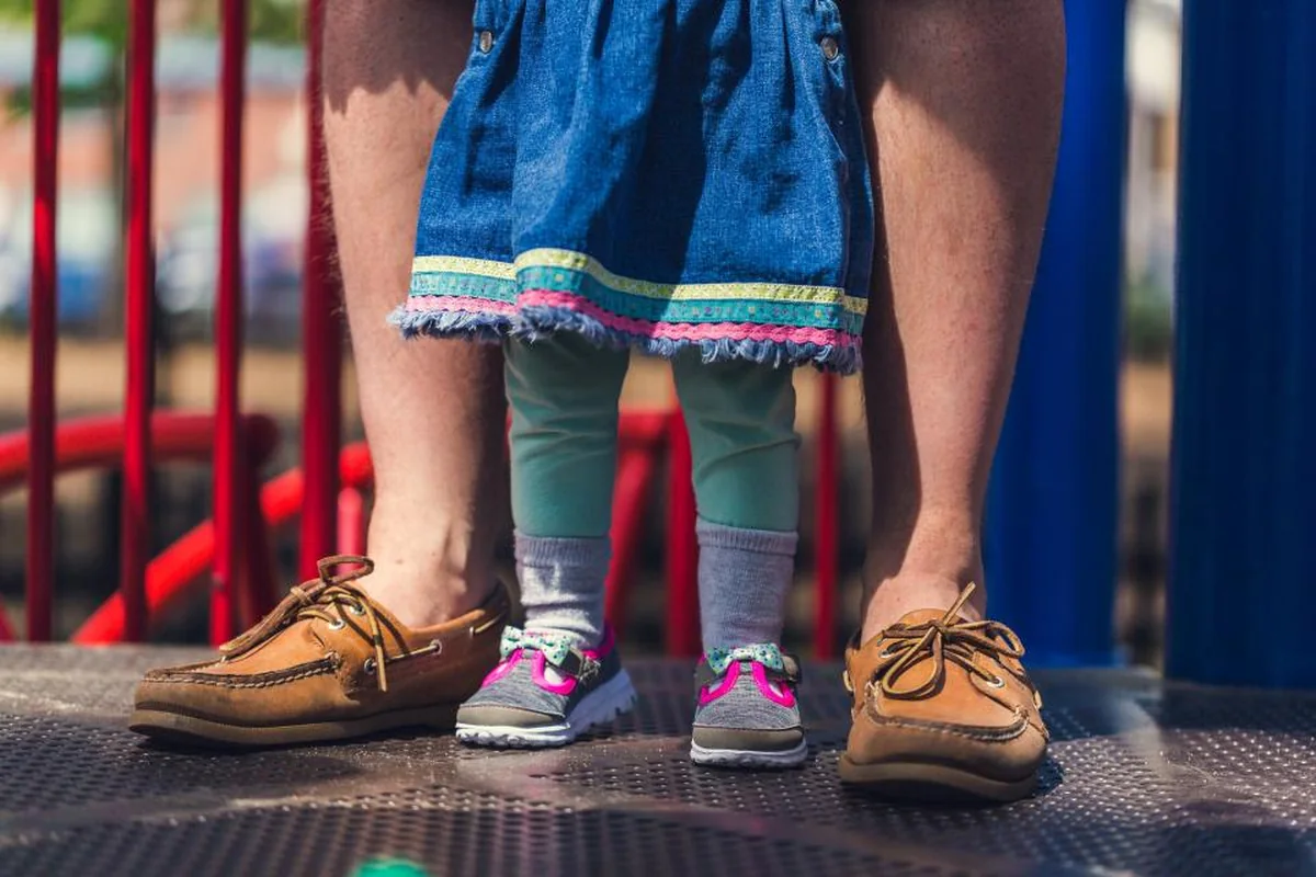 Pies de un adulto con náuticos marrones a cada lado de los pies de una niña con zapatillas y falda vaquera en un parque infantil