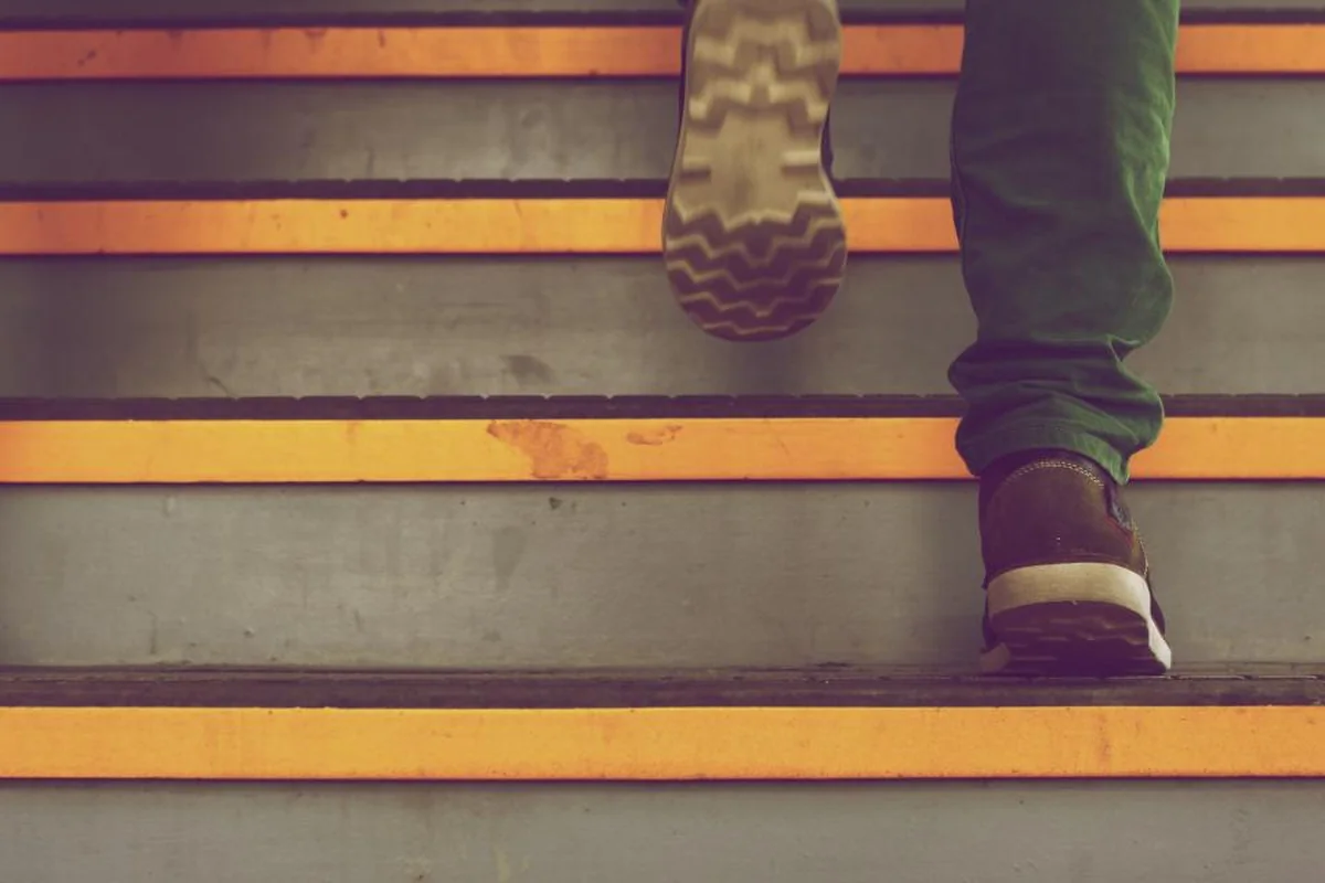 Pies de una persona con pantalón verde y botas marrones subiendo una escalera gris con bandas amarillas en el borde de los escalones