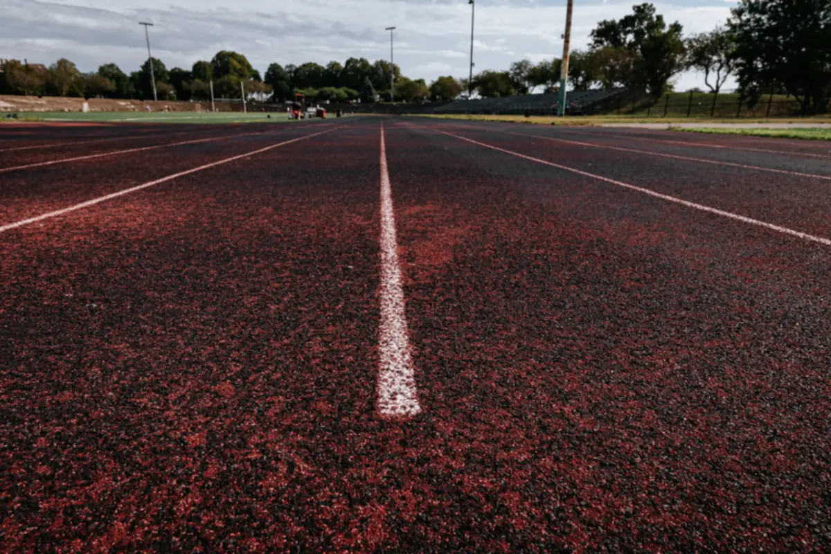 Pista de atletismo vacía de color rojo oscuro vista a ras de suelo, con líneas blancas que separan las calles y árboles al fondo