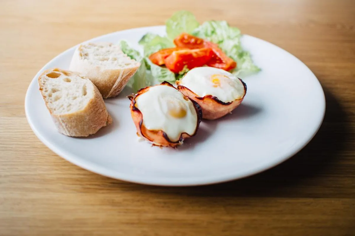 Un plato blanco con dos huevos al horno, trozos de pan y ensalada con tomate sobre una mesa de madera